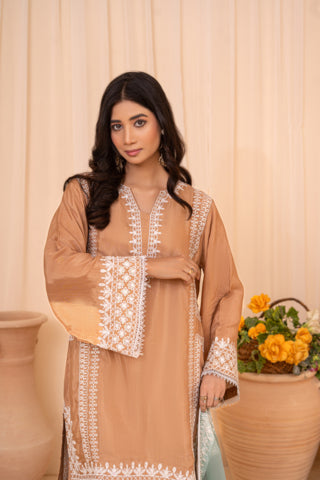 A woman modeling a beige traditional tunic with white embroidered details, standing in front of a cream-colored curtain with decorative terracotta pots and yellow flowers in the background.