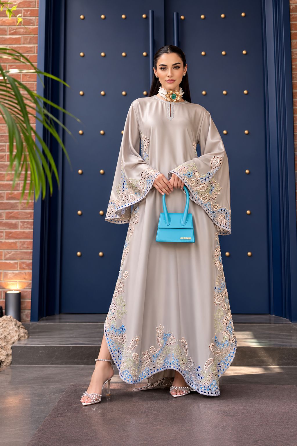 Woman in a long, light-colored embroidered dress holding a blue handbag against a dark blue wall with gold accents.