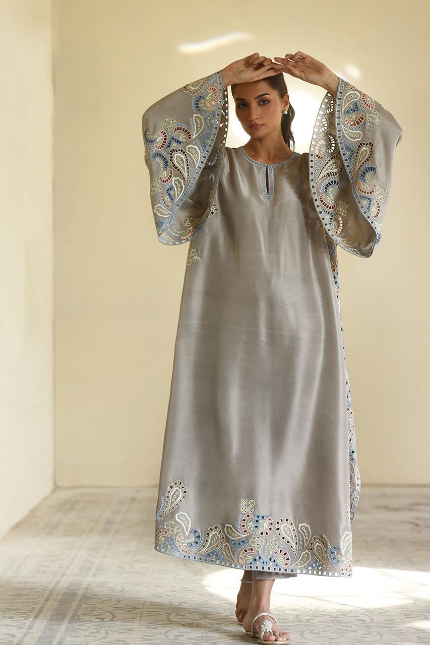 Woman wearing a long, light gray dress with floral embroidery in a bright room.